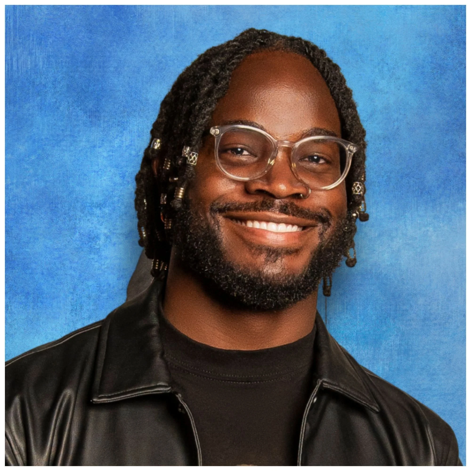 Portrait of a smiling man with glasses, dreadlocks, and a beard, wearing a black leather jacket, against a blue background.