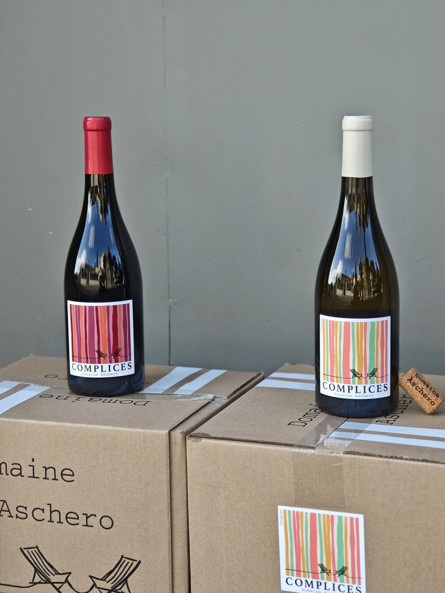 Deux bouteilles de vin avec étiquettes colorées, posées sur des cartons en carton kraft. La bouteille de gauche a une capsule rouge et l'autre une capsule blanche. Au fond, un mur gris.