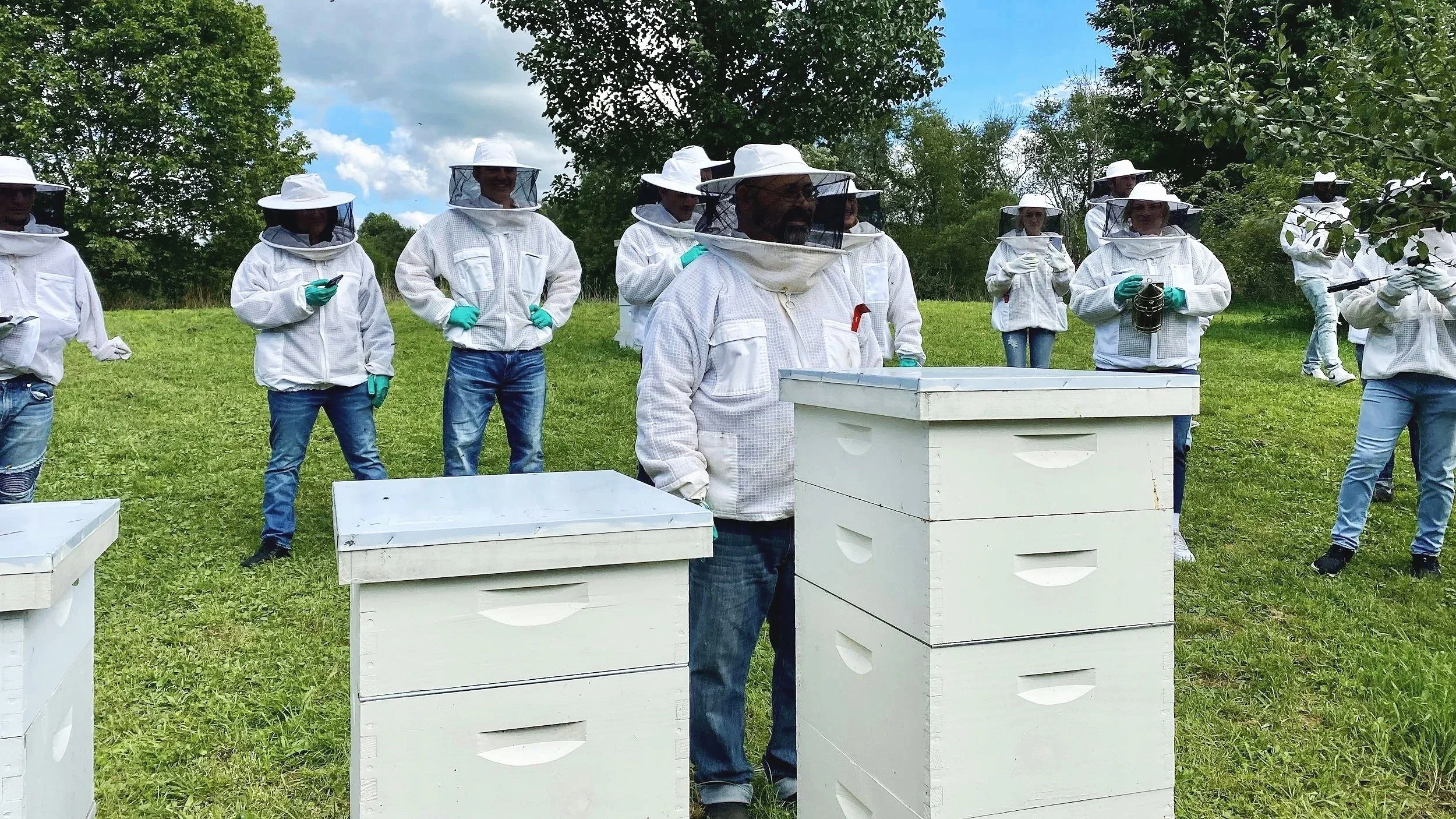 Group of people in beekeeping suits standing outdoors in a grassy area with trees, some using tools or holding equipment, and beehives in the foreground.