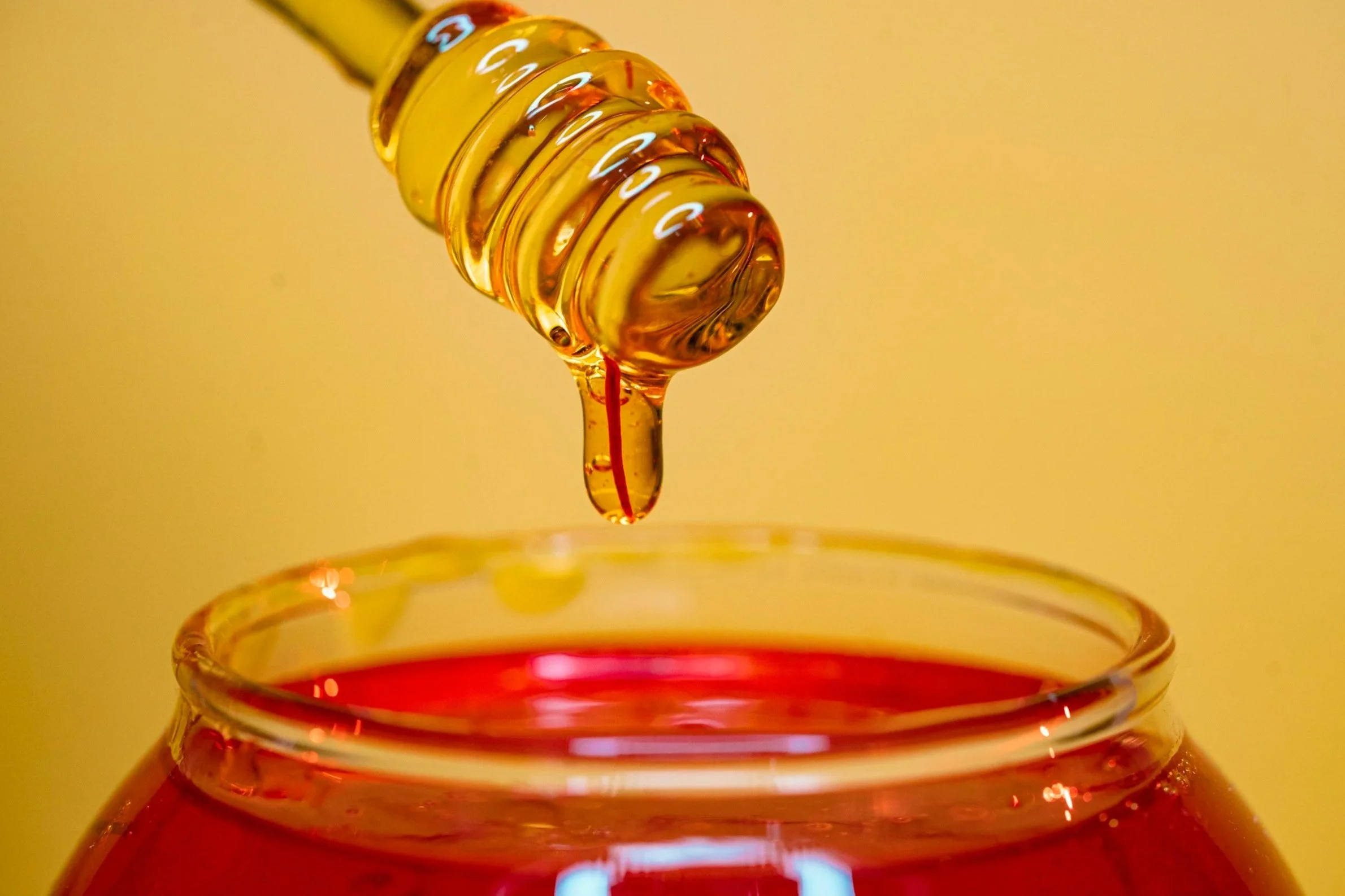 Close-up of a honey dipper releasing honey into a glass jar with red honey inside, against a yellow background.
