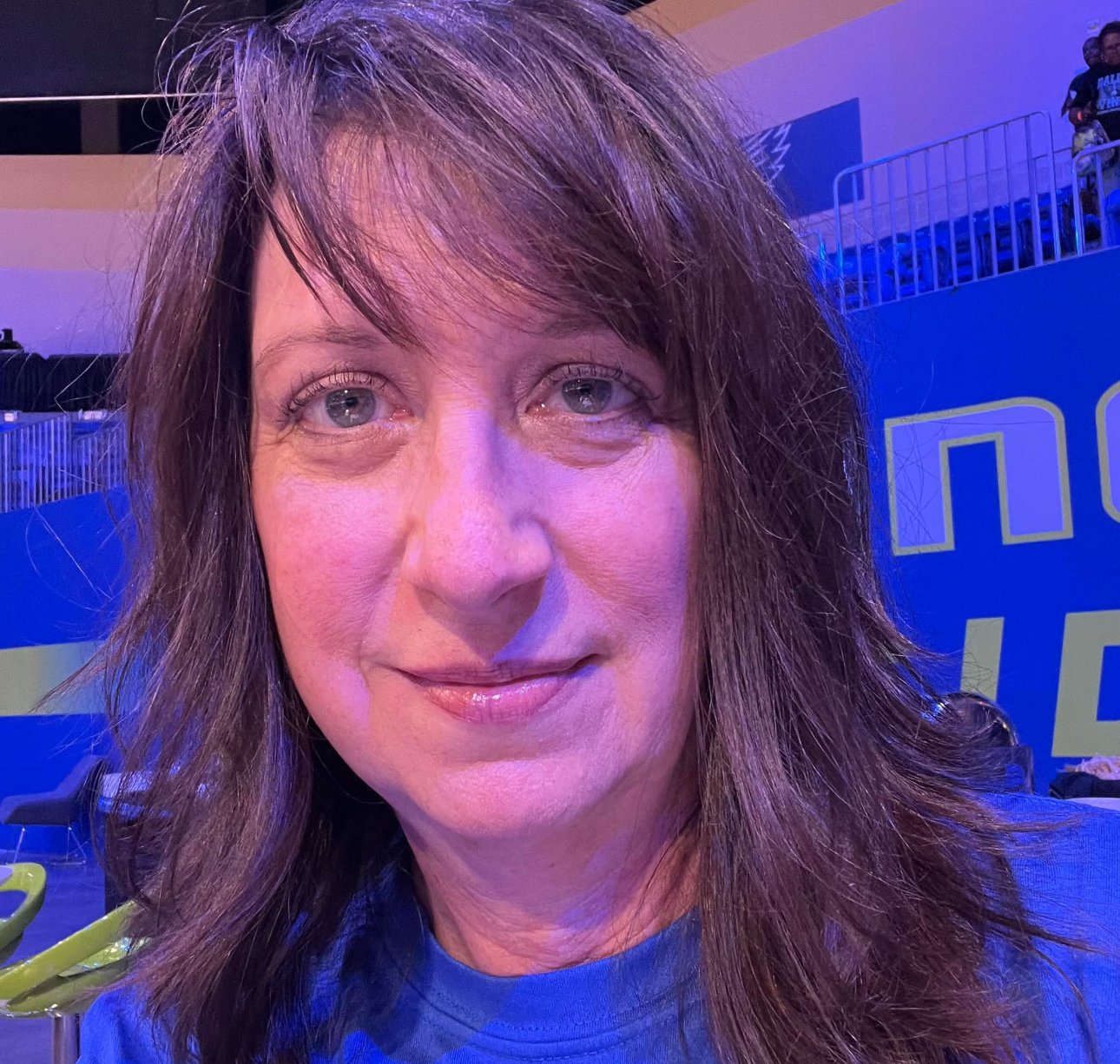 A woman with shoulder-length brown hair and light skin, smiling at the camera, in a setting with blue and purple lighting and stadium seating in the background.
