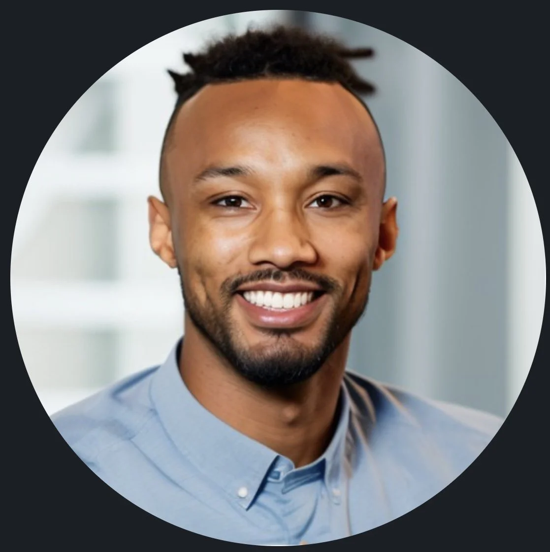 Close-up portrait of a smiling young man with short dreadlocks and a goatee, wearing a light blue button-up shirt, in a professional indoor setting.