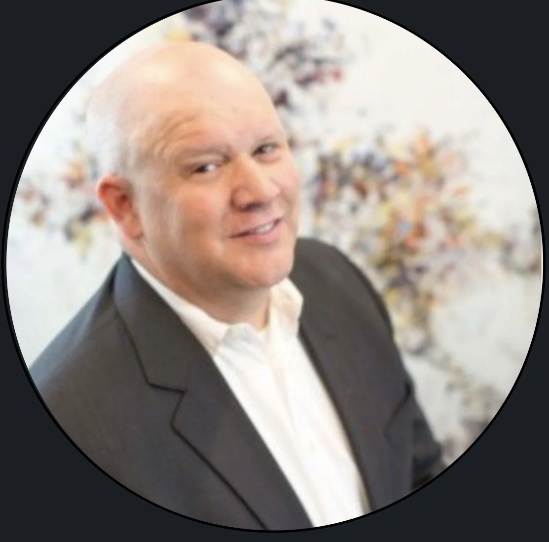 A bald man in a dark suit and white shirt smiling, with a blurred floral background.