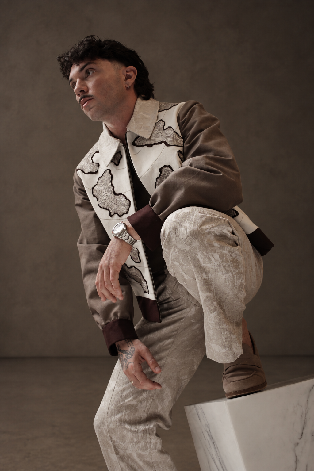 A man wearing a cream and brown jacket with map-like motifs and patterned ivory trousers, with one foot resting on a marble block.