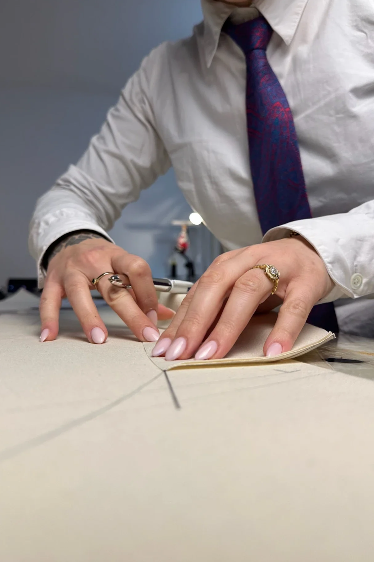 Une personne habillée en blanc avec une cravate violette travaille sur un projet de couture ou de fabrication, les mains posées sur un large morceau de tissu ou de matériau sur une table. La personne porte des bagues, et l’image montre une activité artisanale ou professionnelle.