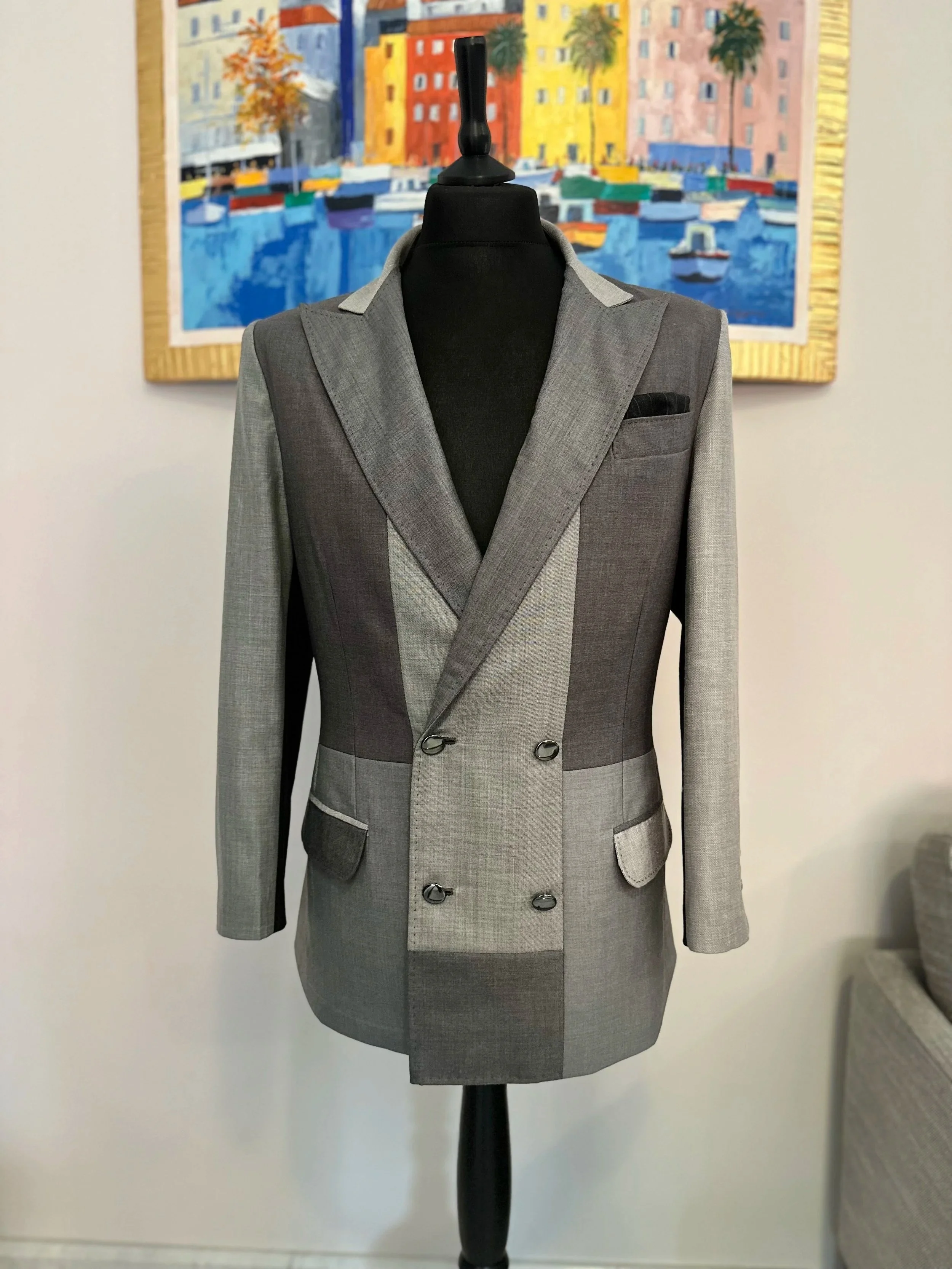 Costume homme en gris avec détails contrastants, manches longues et poche dans la poitrine, exposé sur un mannequin, avec une peinture colorée d'un port avec des bâtiments, des palmiers et des bateaux en arrière-plan.