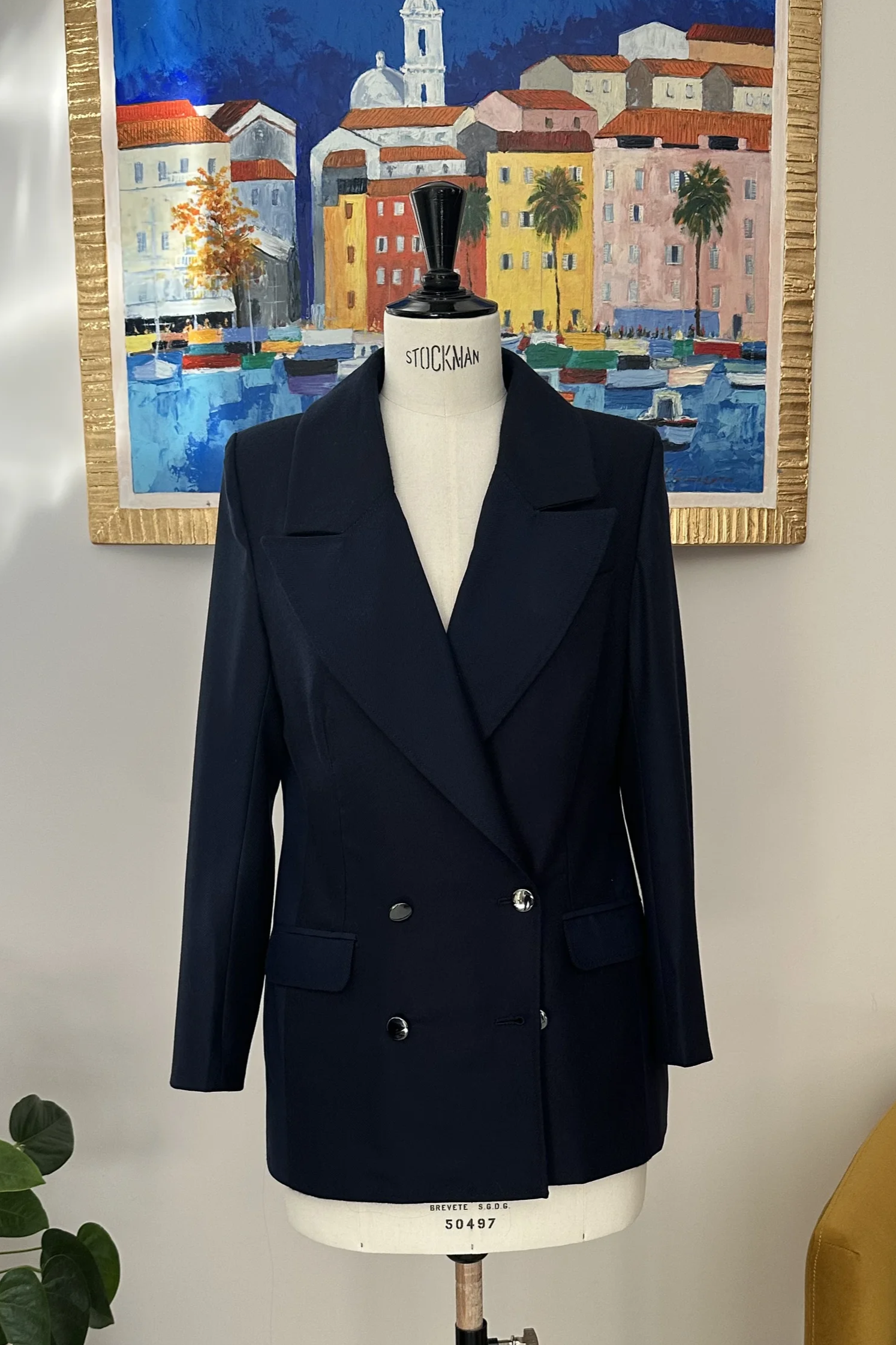 Vitrine avec un mannequin portant un blazer noir, une peinture représentant un port coloré avec des maisons et des palmiers, un fauteuil jaune avec un coussin beige, et une plante verte dans un pot blanc.