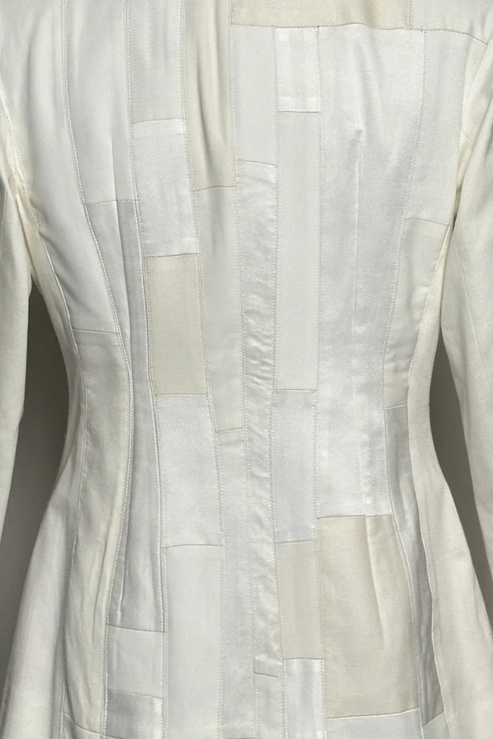 Mannequin portant un blazer blanc devant une peinture colorée représentant un port avec des bateaux et des bâtiments aux couleurs vives.