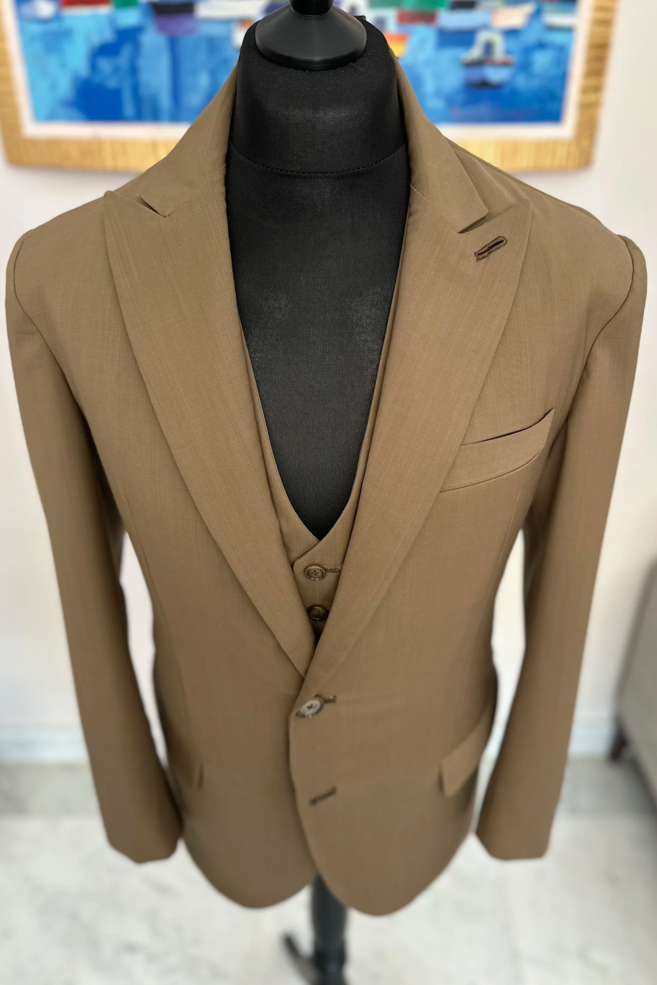 Mannequin portant un costume beige avec une chemise ou un t-shirt gris foncé, derrière un tableau avec des bateaux colorés.