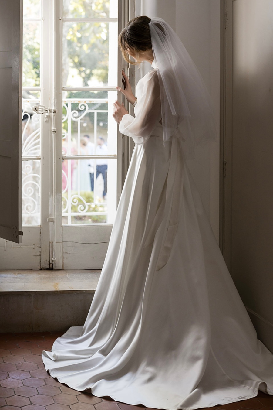 Une mariée en robe blanche et voile regarde par une fenêtre ouverte lors d'un mariage