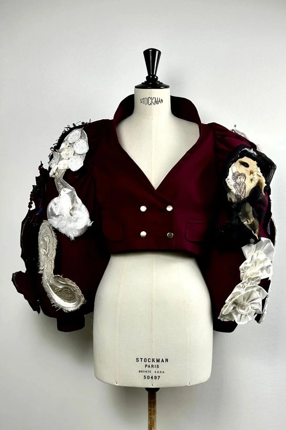 Un mannequin d'épaule portant une veste rouge avec des décorations en tissu blanc, noir, et beige, exposée contre un mur blanc.
