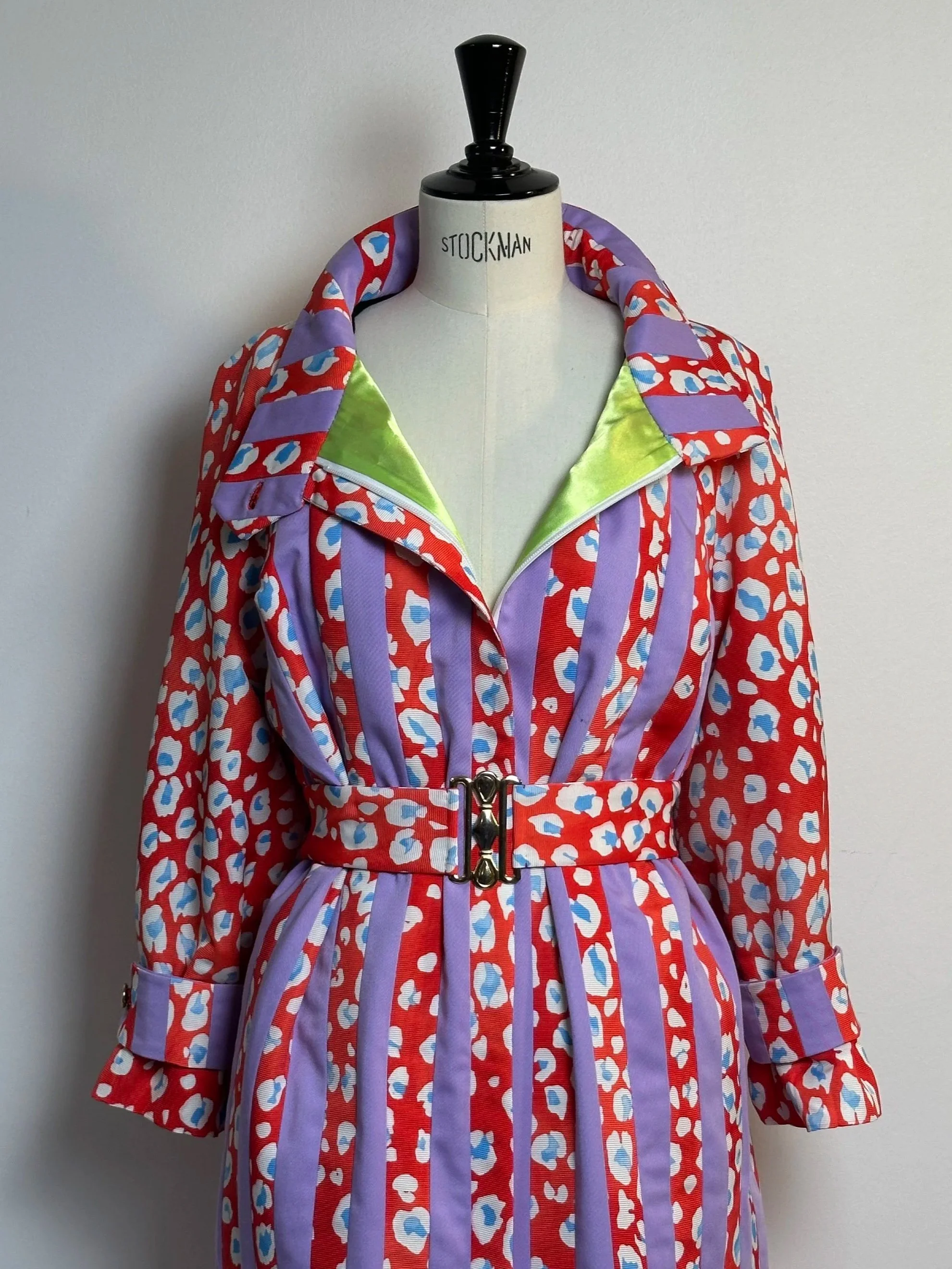 Mannequin portant une robe longue à motifs rouges, violets et blancs avec des rayures violettes et des accents verts à l'ouverture du col, avec une ceinture argentée, devant un mur blanc.