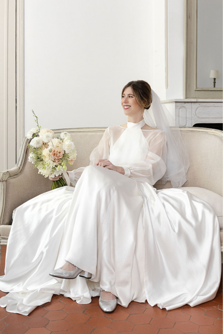 Une femme en robe de mariage blanche assise sur un canapé beige, tenant un bouquet de fleurs, dans une pièce élégante avec un miroir et une cheminée.