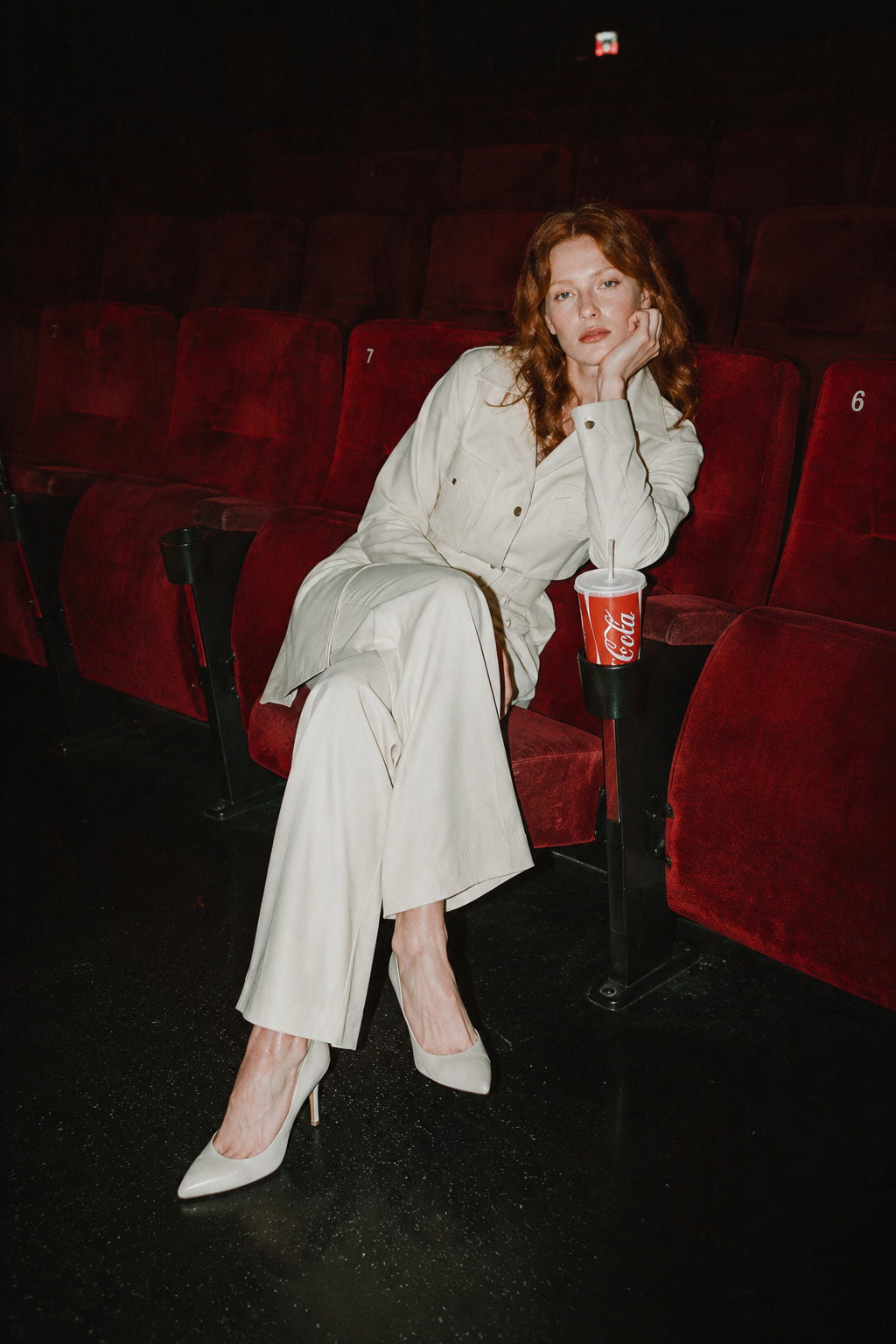 Femme aux cheveux roux assise dans une salle de cinéma, portant une tenue claire, avec une boisson Coca-Cola à ses côtés.