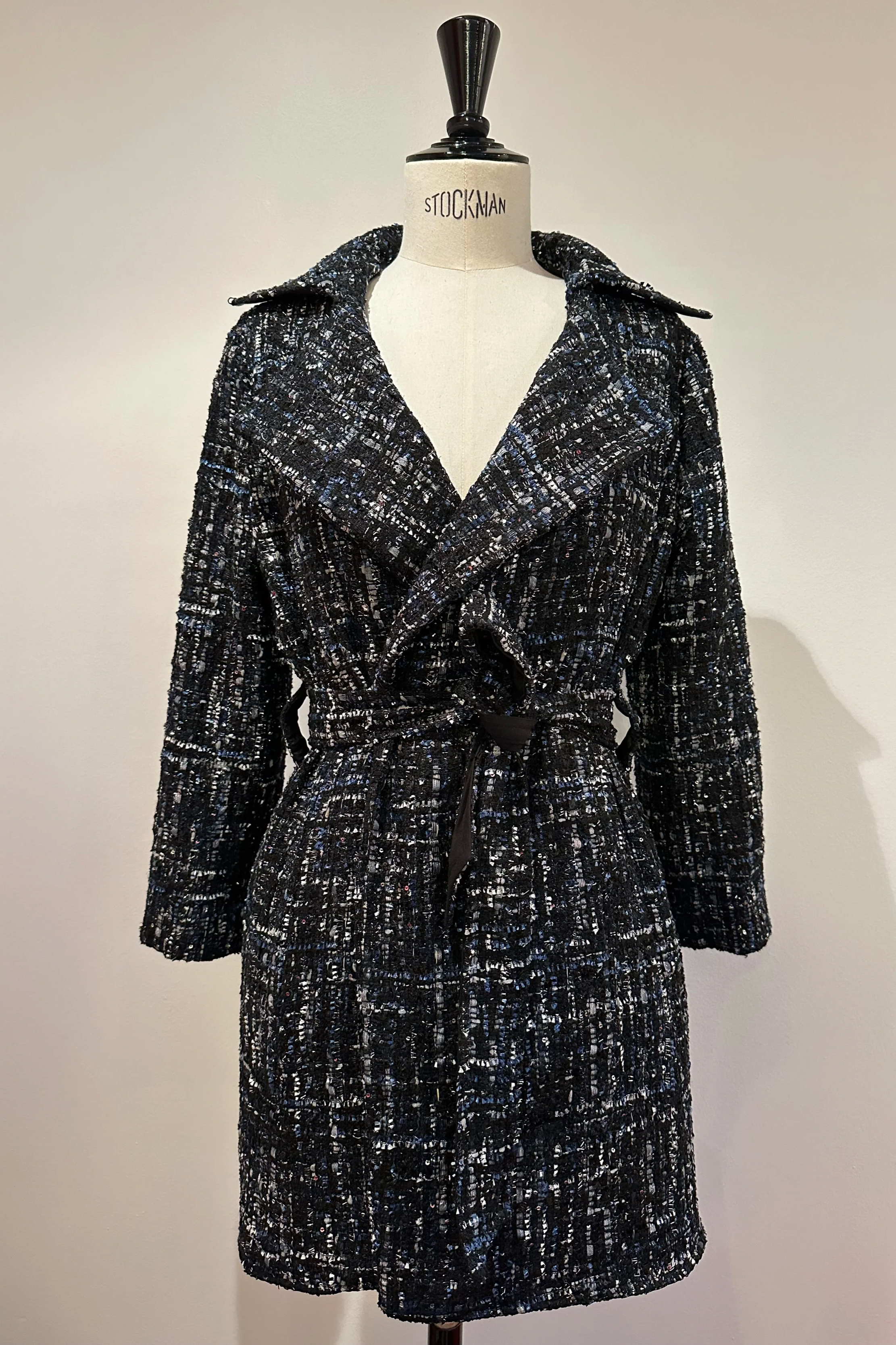 Mannequin portant un manteau long avec une couture en tweed noir, blanc et gris, avec une ceinture à la taille, présenté sur un fond neutre.