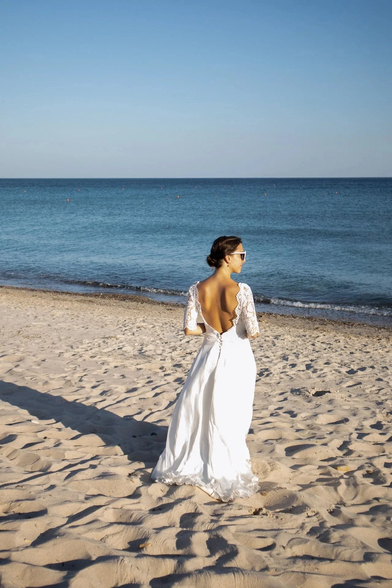 Une femme en robe de mariage blanche se tient sur la plage, regardant vers la mer, portant des lunettes de soleil, avec des dunes de sable au premier plan et l'océan en arrière-plan.