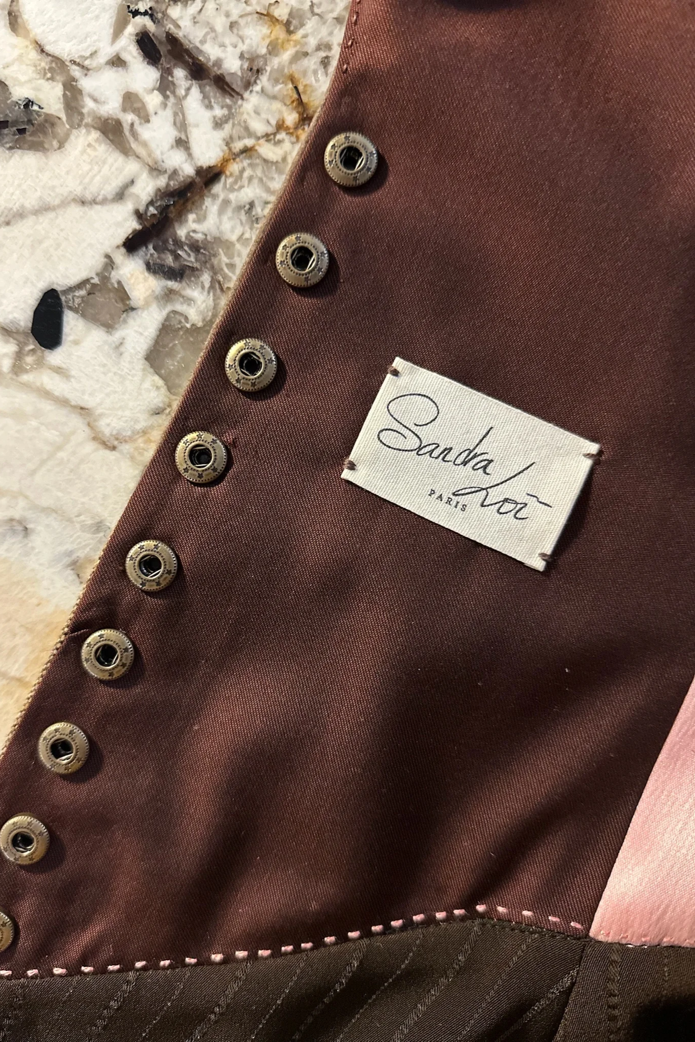 Détail d'une veste avec des boutons ronds en métal, une étiquette blanche avec l'écriture 'Sanda Lou PARIS', et un fond de pierre blanche et foncée.