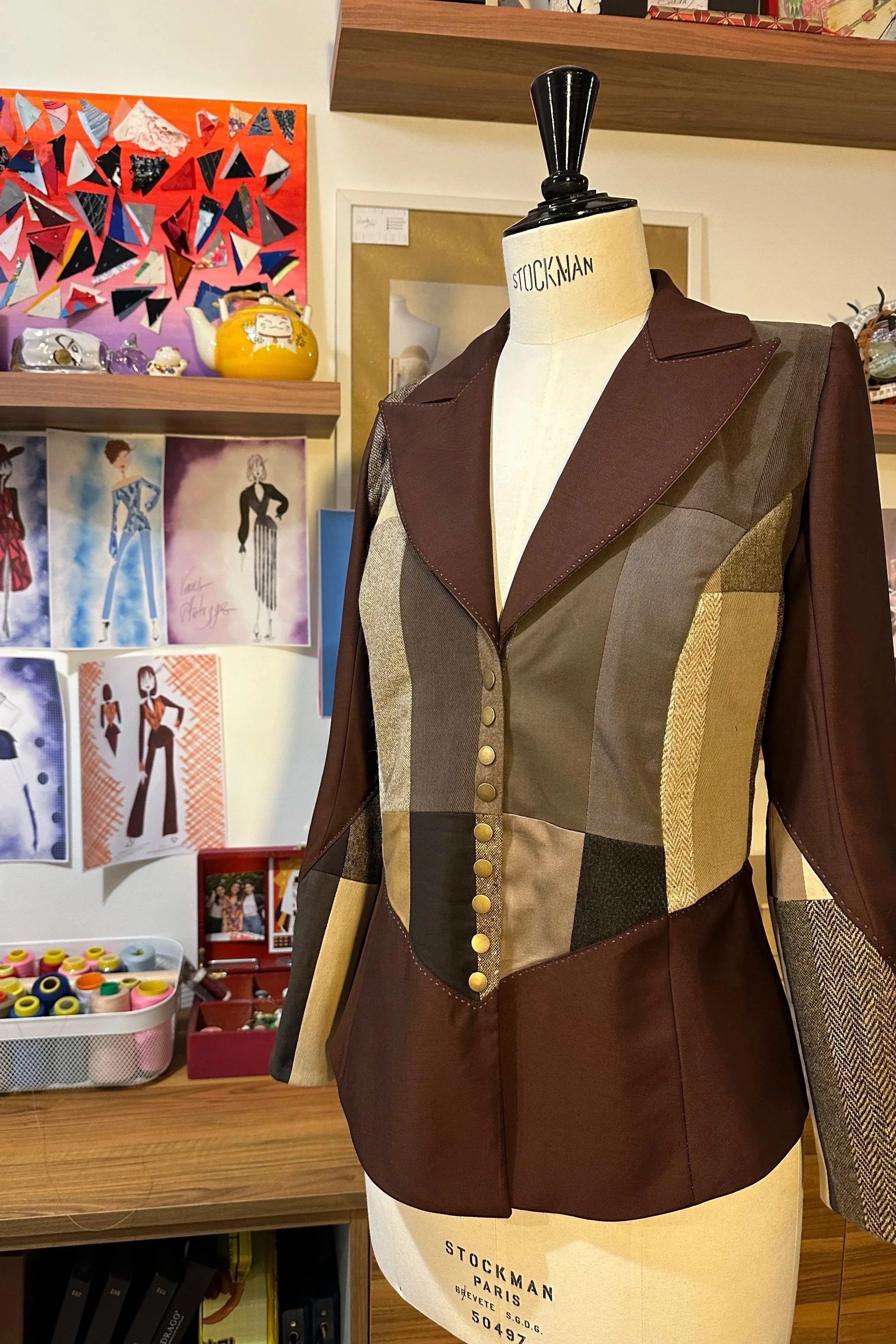 Mannequin portant un tailleur en patchwork marron, beige et à motifs écossais, dans un magasin de mode. En arrière-plan, affiche avec des illustrations de modèles de vêtements et diverses objets de décoration.