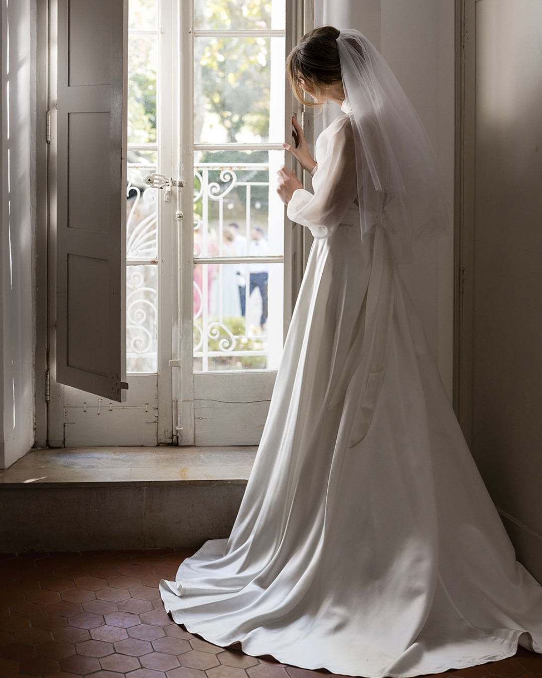 A bride in a long ivory gown with sheer sleeves and a veil, standing beside an open window.