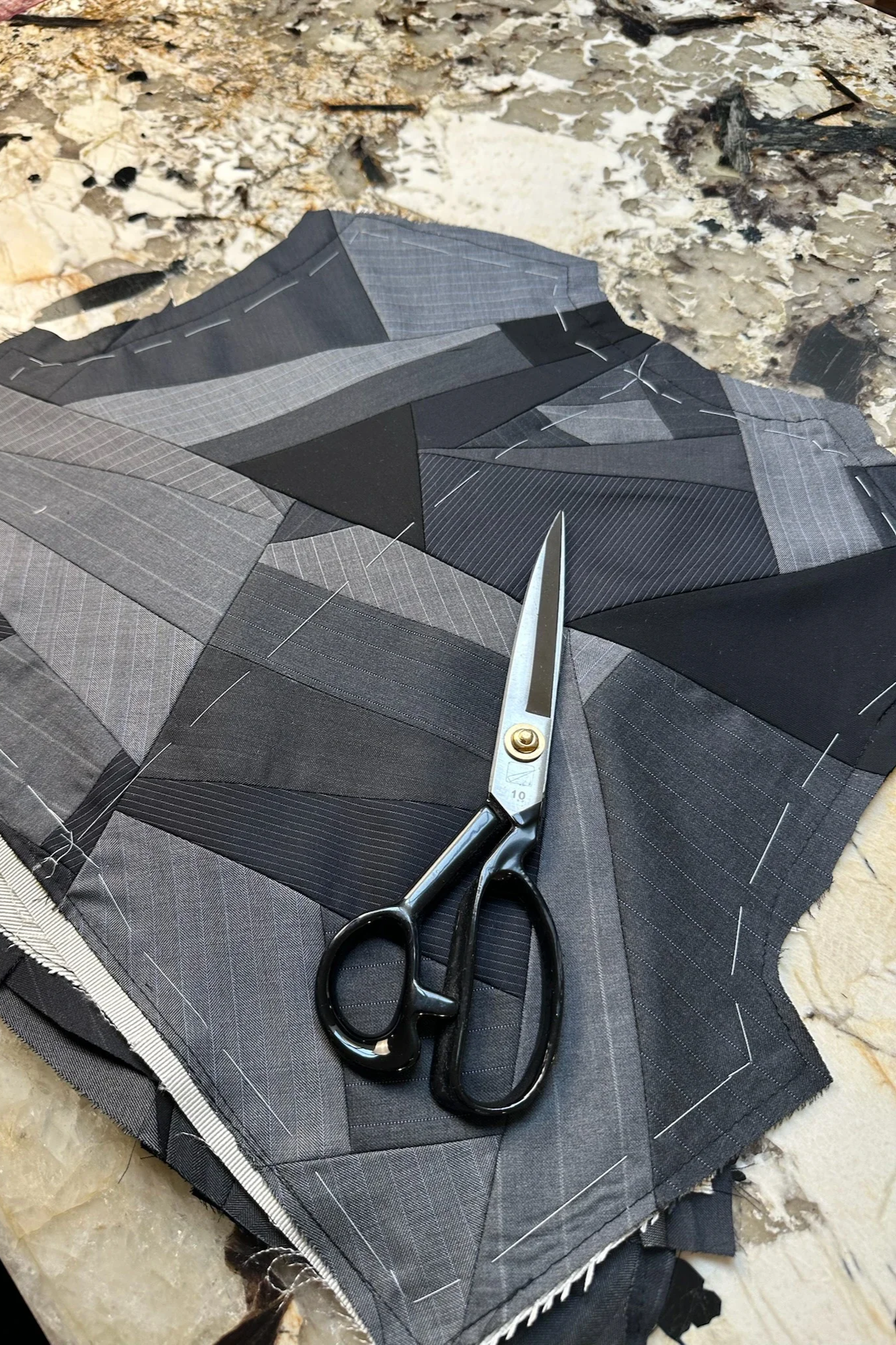 Un tissu à carreaux gris est étalé sur une surface, avec une paire de ciseaux à manches noires posés dessus. Le tissu est en cours de couture avec des épingles en place.