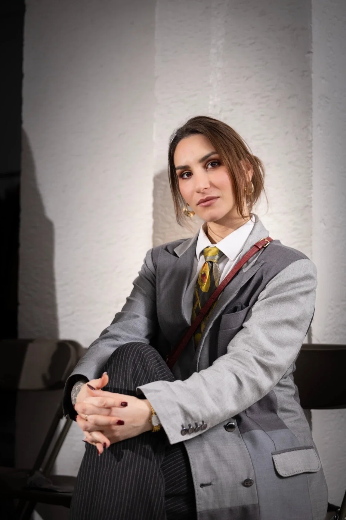 Femme assise en costume professionnel gris, portant une chemise blanche, une cravate avec un motif, et des boucles d'oreilles en or, assise en face d'un mur blanc avec un regard sérieux.