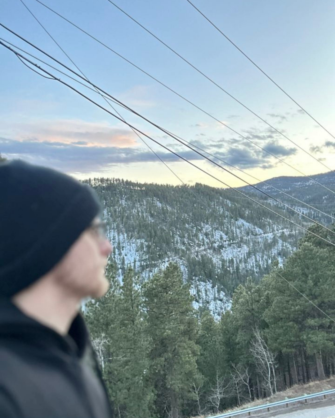 A blurry person wearing a black hoodie and black hat in the foreground, with a mountain landscape, pine trees, snow patches on the mountain, and power lines in the background during late afternoon or early evening.