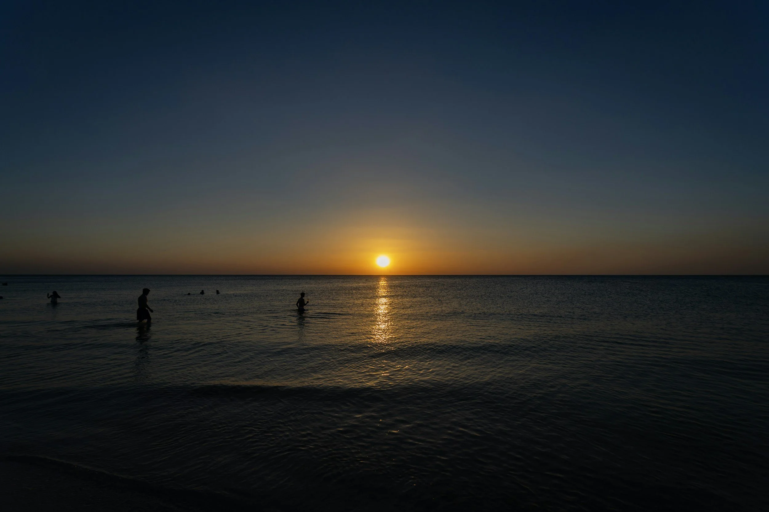 People wading in the ocean at sunset with the sky transitioning from blue to orange.