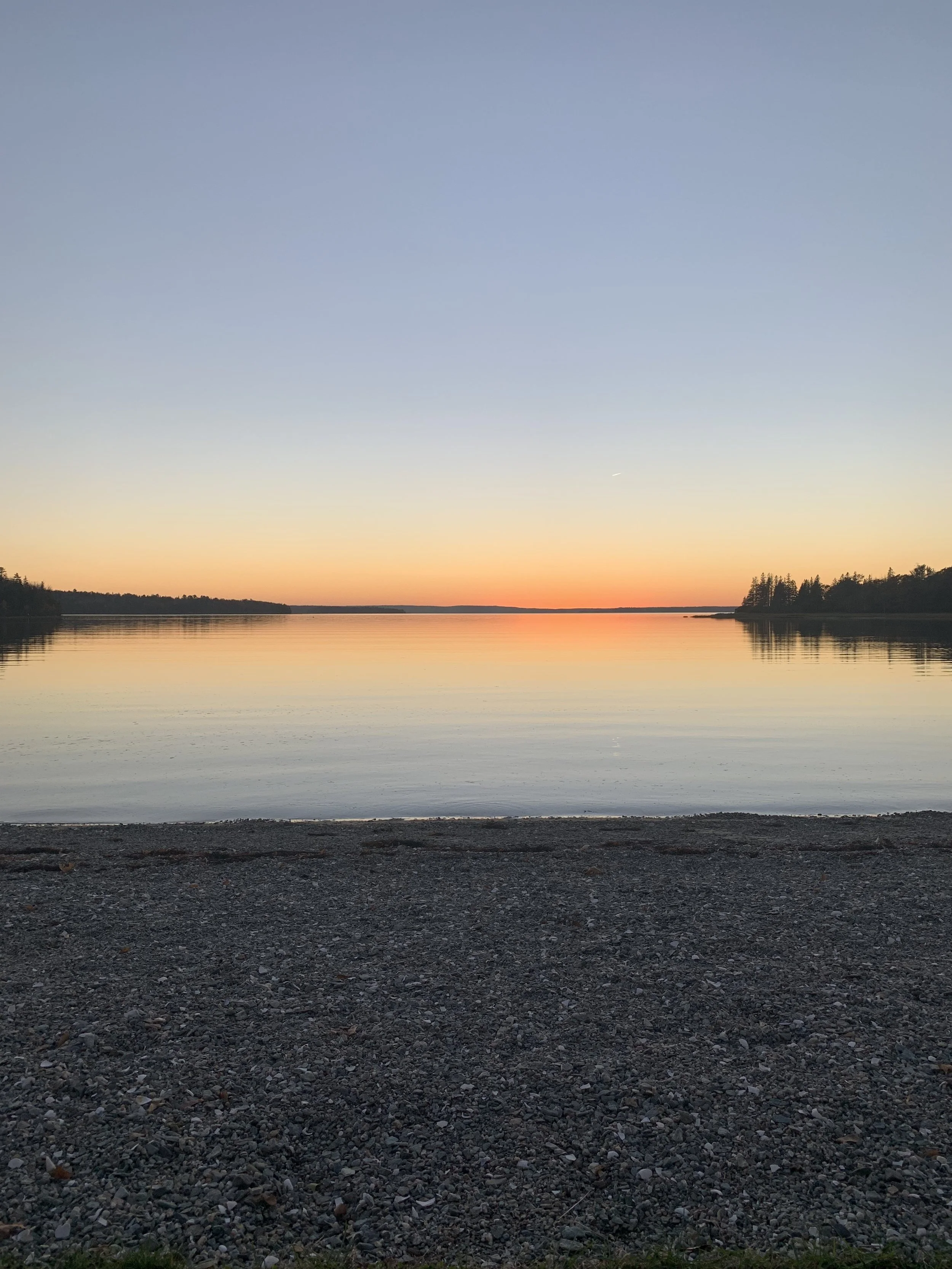 Sunset over a calm body of water with a gravel beach in the foreground and distant forested shoreline.