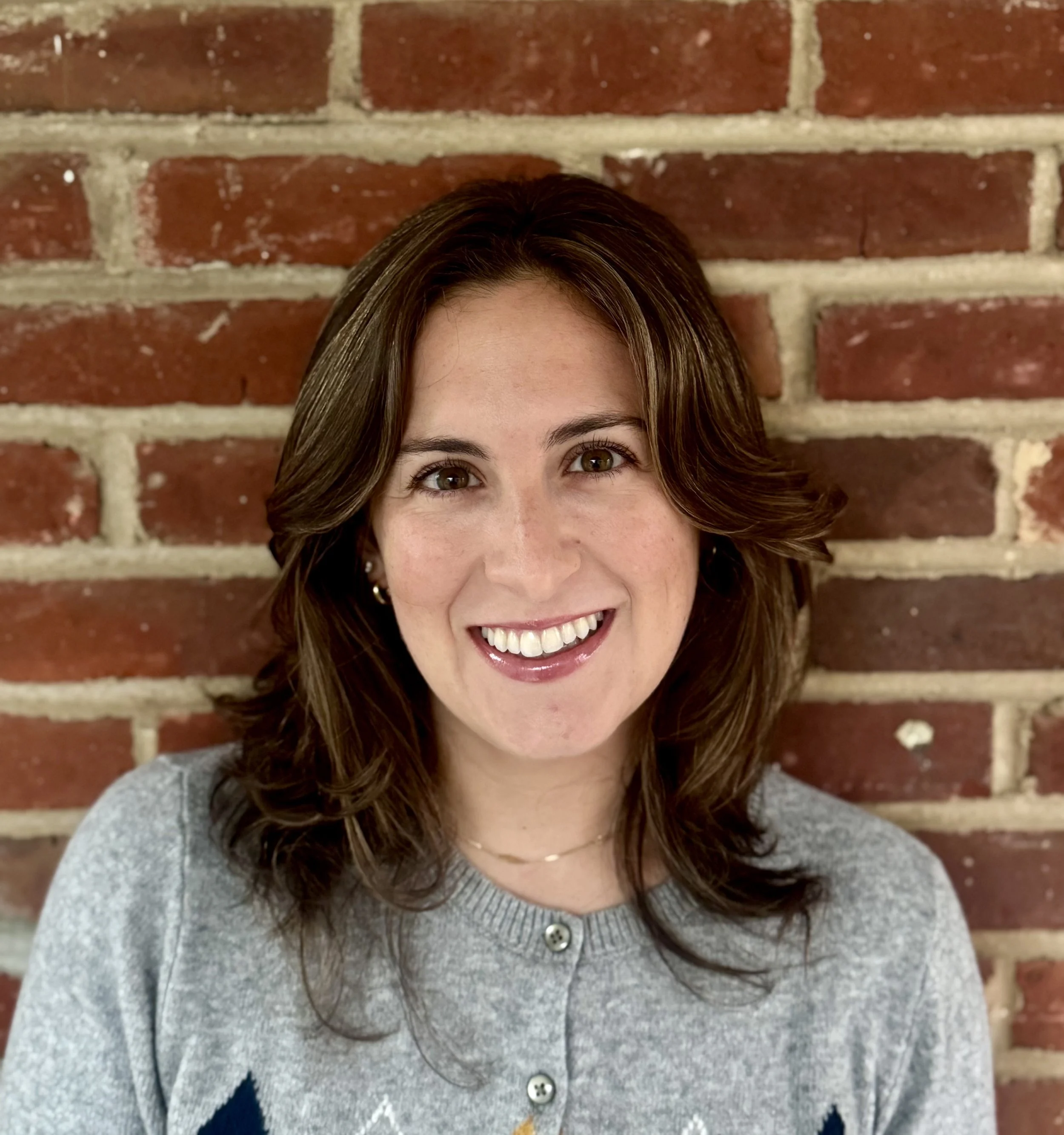 Headshot of smiling woman with brown hair and light skin, standing against a brick wall.