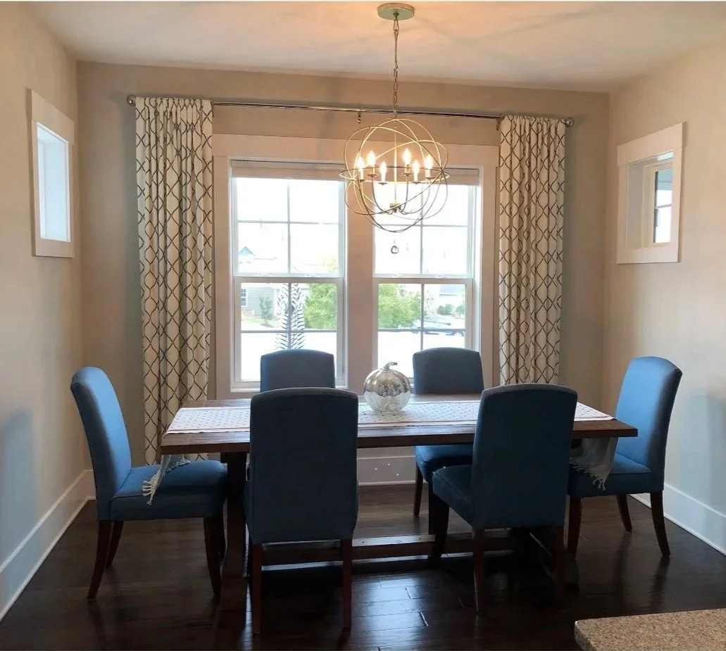 Before: a builder-standard dining room with beige walls, blue upholstered chairs, a farmhouse table, and an orb chandelier over patterned floor-length curtains.