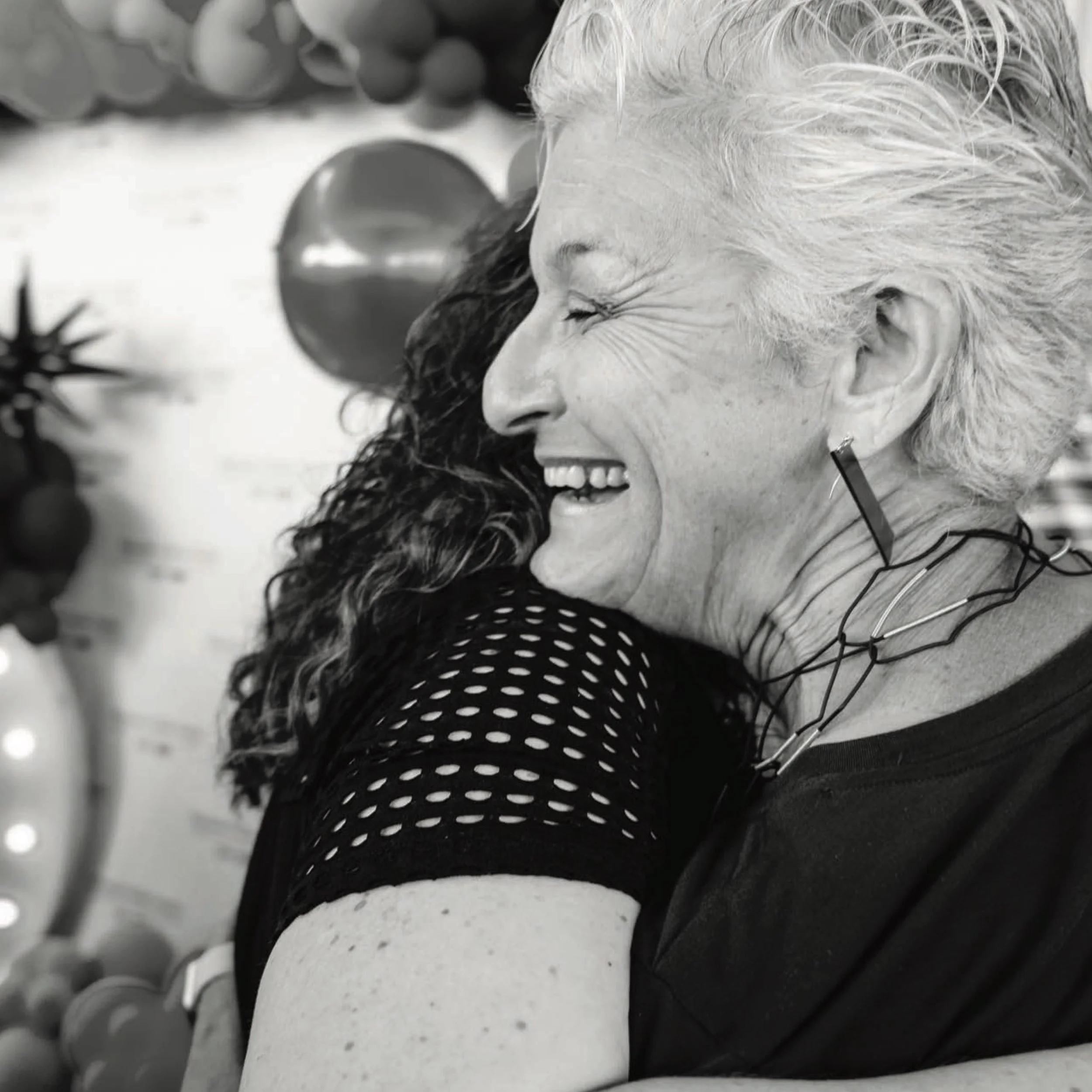 Black and white photograph of two guests embracing and laughing in front of a balloon display at the Houston Matters tenth anniversary party.