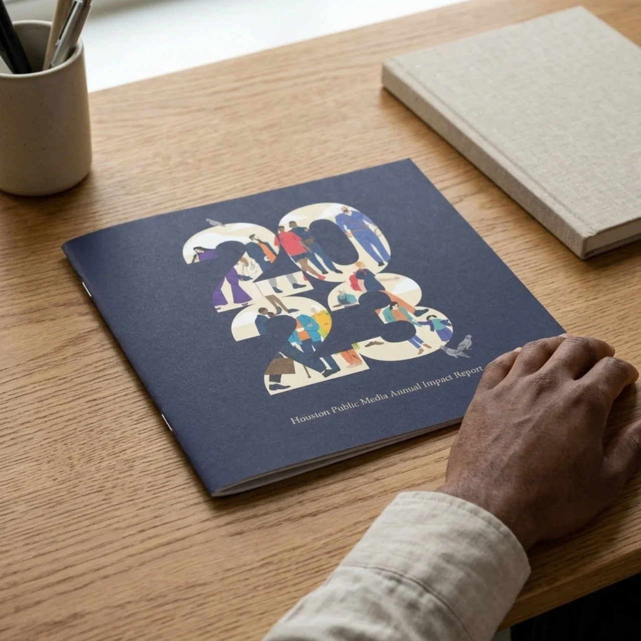 The 2023 Houston Public Media Annual Impact Report on a wood desk, its navy cover carrying illustrated figures cut out of the numerals 2023.