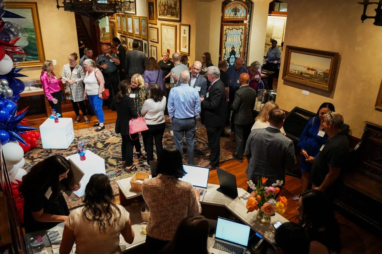 Guests fill a wood-paneled gallery at the Bryan Museum during Houston Public Media's Galveston County News Bureau launch reception, with a red-white-and-blue balloon display at the entrance.