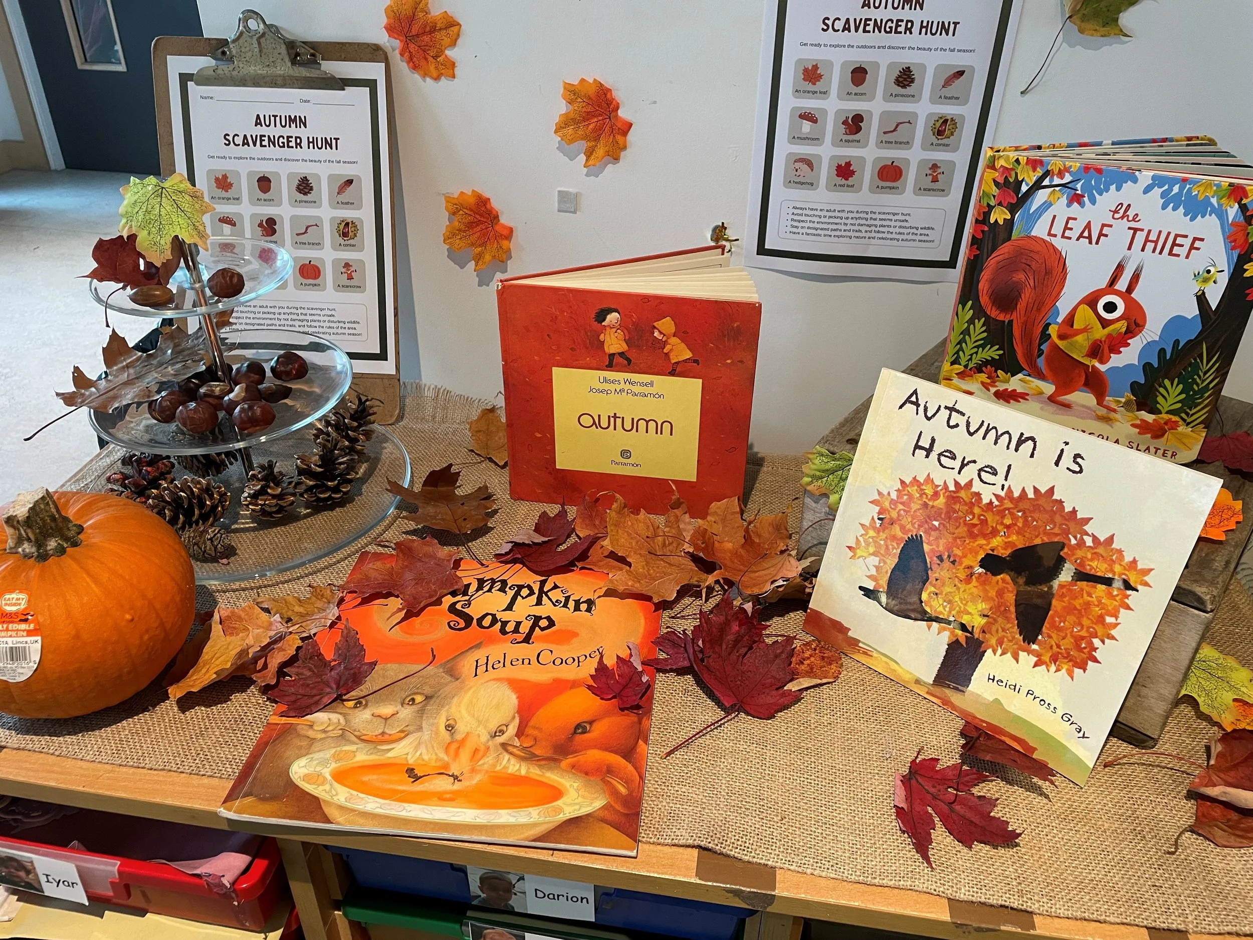 Display table with fall-themed books, pumpkins, and decorative leaves for autumn. Sign mentions autumn as the season, and there are posters about an autumn scavenger hunt.