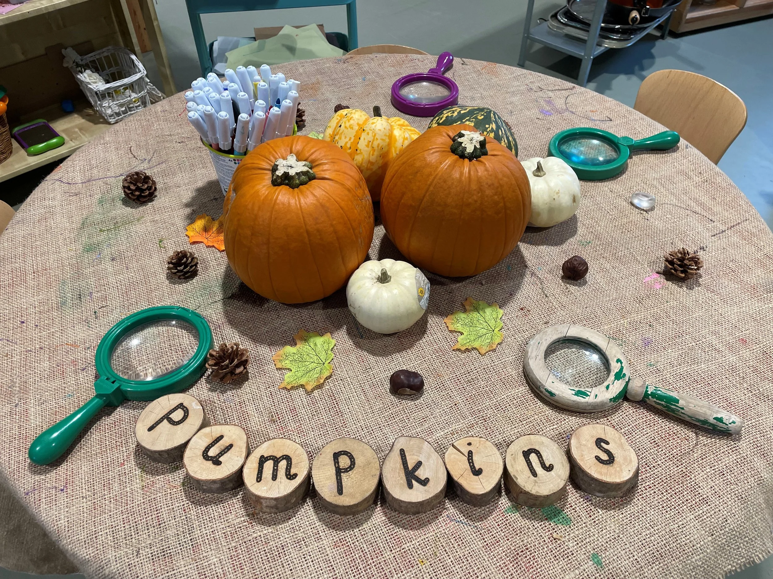 A table decorated with autumn pumpkins, leaves, and pinecones, with a container of markers and small magnifying glasses. The word "pumpkins" is spelled out with wooden letter tiles.