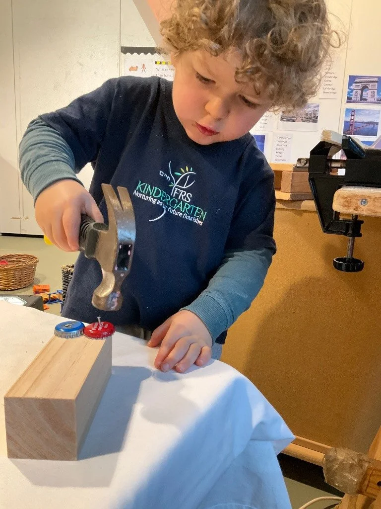 A young boy with curly hair using a hammer to nail a bottle cap onto a wooden block in a classroom setting.
