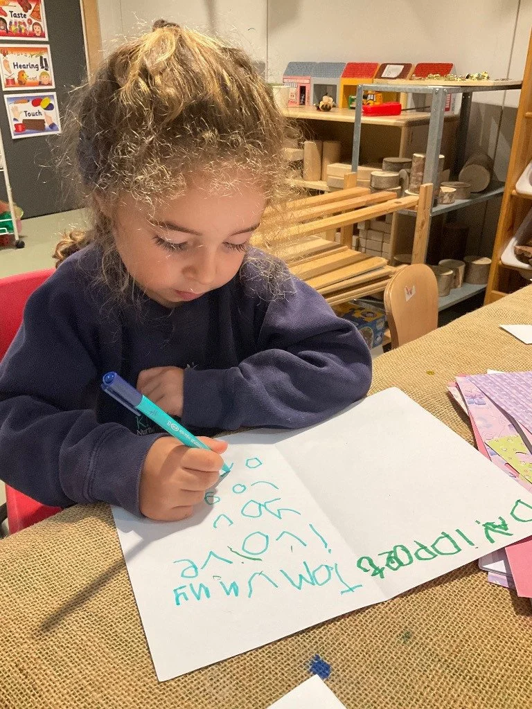 A child with curly hair writing on a large white sheet of paper at a table in a classroom. Nursery classroom. Finchley, London