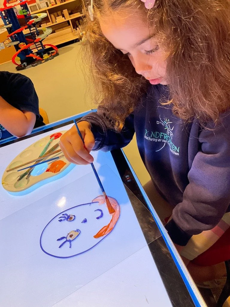 A young girl with curly hair painting a smiley face on a digital drawing screen, using orange and blue paints. She is wearing a dark sweatshirt with a logo that says 'KINDER' and the background shows a classroom setting with toys and shelves.