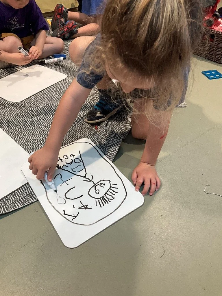A young girl with curly hair drawing on a whiteboard with a black marker, surrounded by other children sitting on a rug.