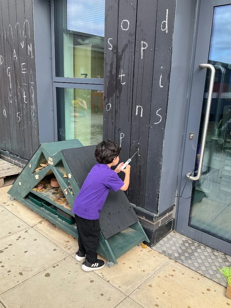 A young boy in a purple shirt and black pants writing on a black wooden wall outside a building with chalk or white marker, with partially visible letters and words like 'ands' and 'Lights' and an accessible door with a glass window nearby.