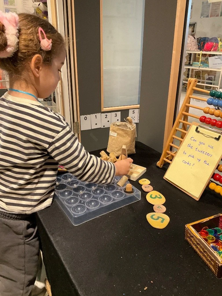 A young girl with a striped shirt and pink hair clips plays a game involving tokens and numbered corks at a children's activity table.
