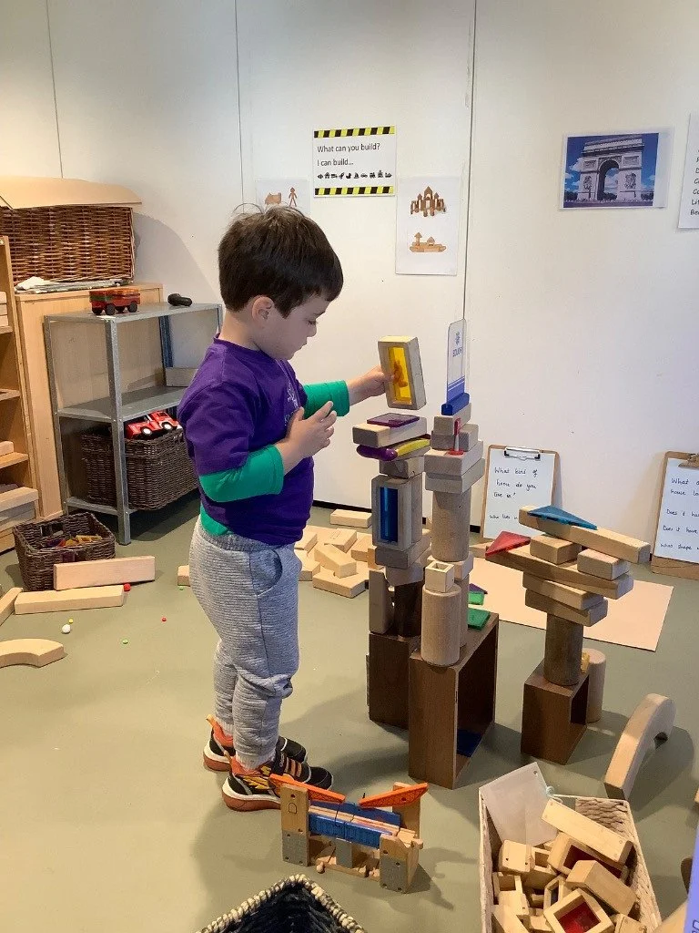 A young boy in gray sweatpants and a purple shirt with green accents plays with large wooden blocks on the floor in a playroom.