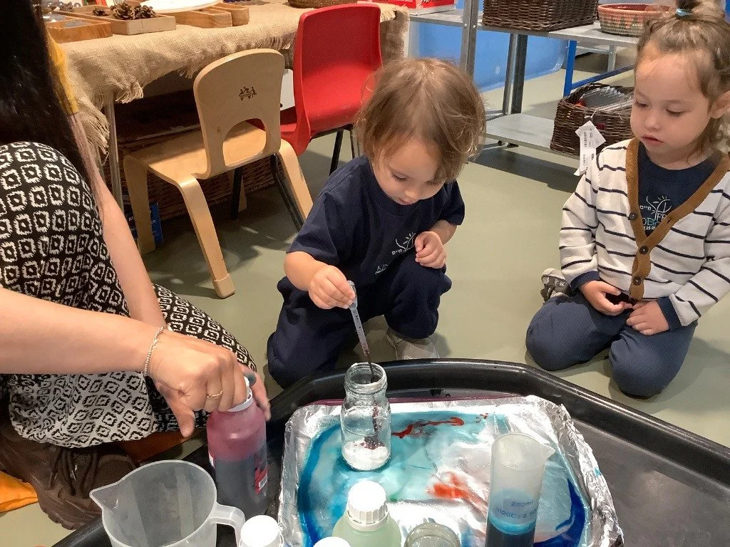 Two young children watching a woman conduct a science experiment with colorful liquids and a small glass jar.