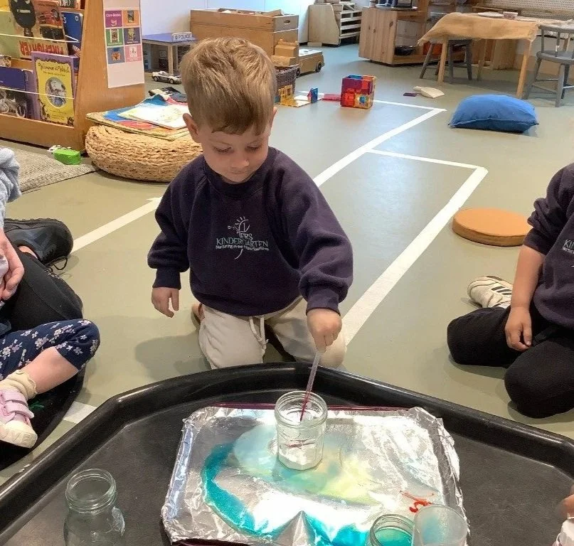 A young boy in a preschool uniform is kneeling on the carpet, observing a science experiment with a jar on a tray covered with aluminum foil, surrounded by children sitting cross-legged.