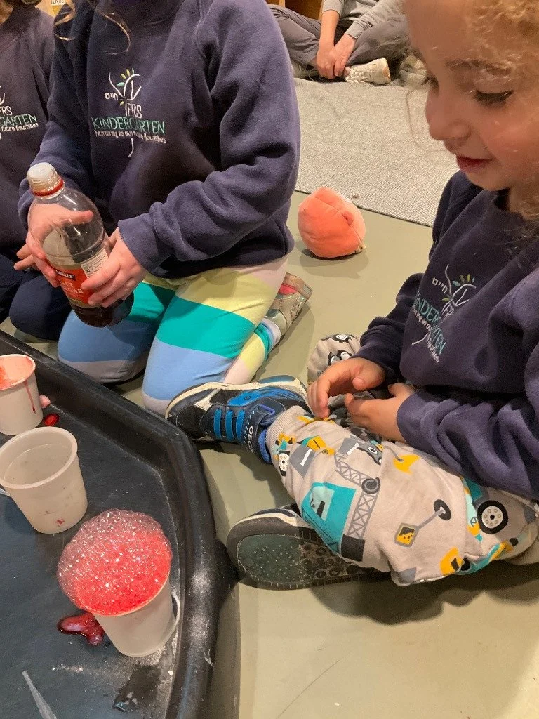 Children sitting on the floor engaged in a science experiment with cups and a tray, one cup filled with red liquid and covered with foam, in a classroom setting.