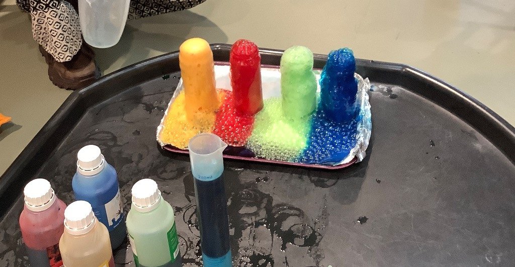 Four bottles of liquid paint in pink, blue, beige, and green, with a test tube containing blue liquid, and a tray with four cylindrical objects sprinkled with different colors of beads—yellow, red, green, and blue.