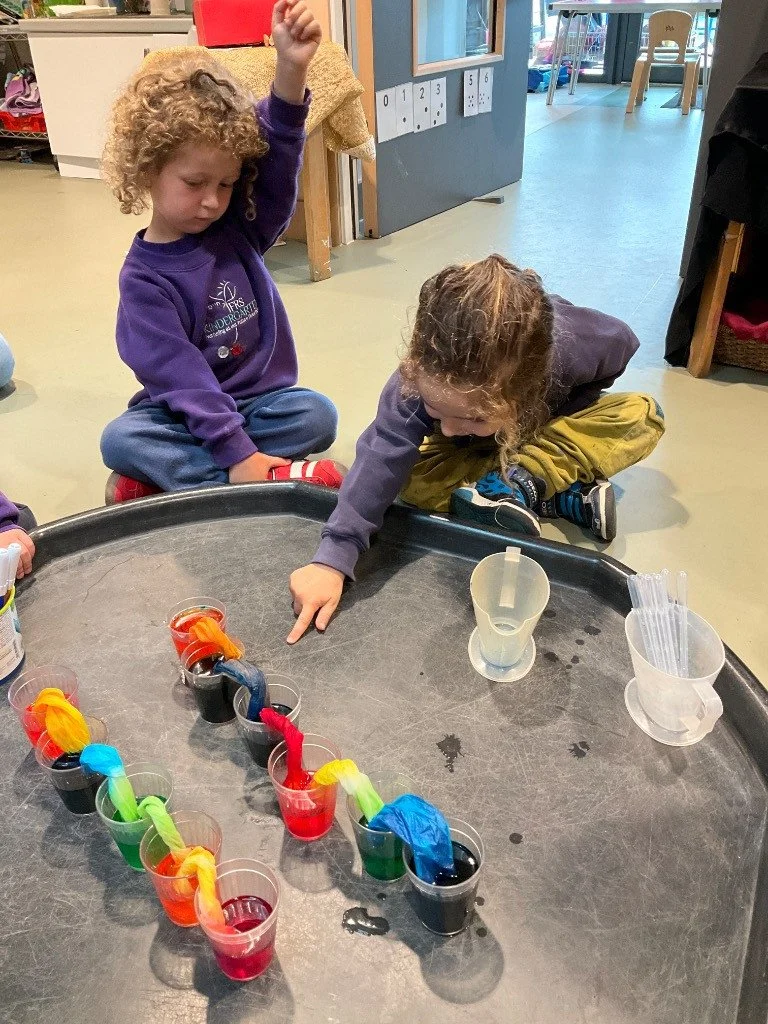 Children participating in a science activity with colored liquids and tissue paper in cups at a table in a classroom.