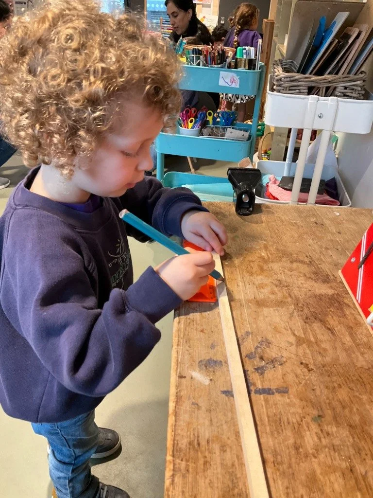 A young child with curly hair wearing a navy sweatshirt and blue jeans is drawing or coloring on a small white paper or notebook on a wooden worktable with a blue marker. There are art supplies and people in the background inside an indoor space.