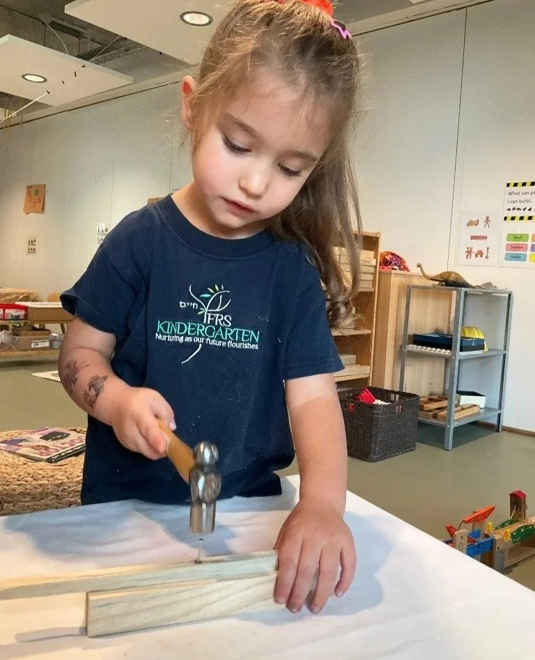 A young girl with a ponytail, wearing a navy T-shirt with the text 'IFRS Kindergarten' and an illustration of a sunrise, is hammering a nail into a wooden piece at a table in a classroom setting.