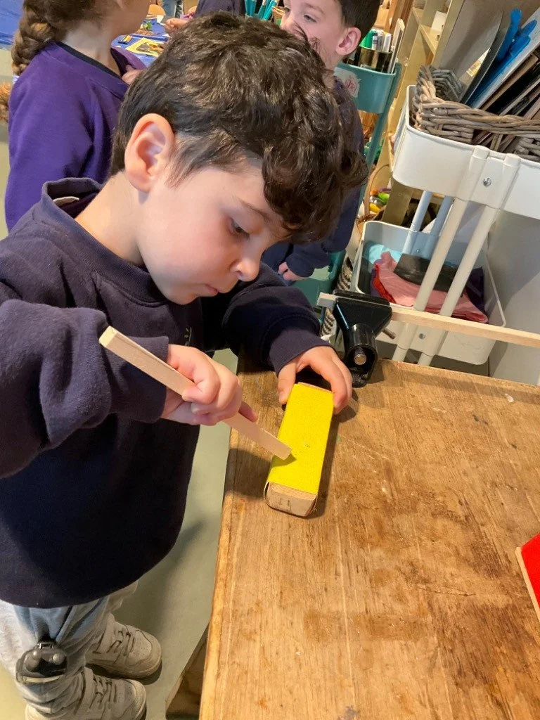A young boy with dark curly hair is using a hammer and a chisel to carve a yellow-painted piece of wood on a worktable in a classroom or workshop setting. Other children are visible in the background.
